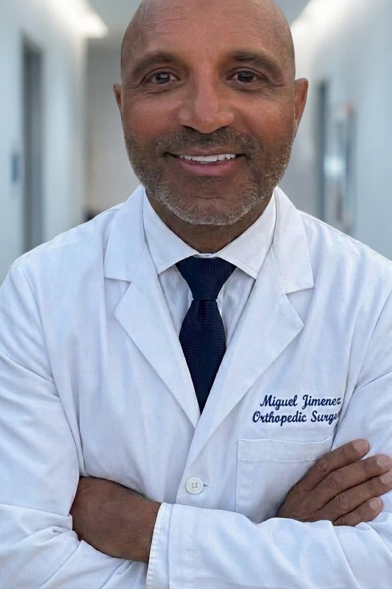 Dr. Miguel Jimenez, arms folded, in a white coat embroidered “Miguel Jimenez, Orthopedic Surgeon”, standing in a clinic corridor.